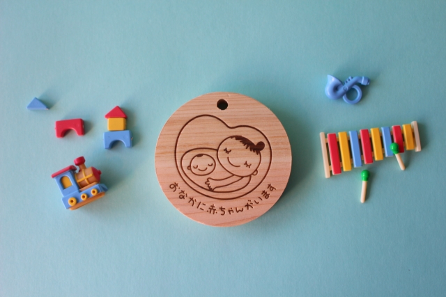 木製のマドレーヌタグに親子のイラストと「おなかに赤ちゃんがいます」と書かれている。周囲にはカラフルなおもちゃや楽器が散らばっている。
